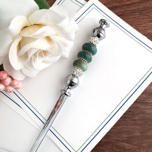 May include: A silver letter opener with a green and white beaded handle. The handle has a silver cap on the top and a silver base at the bottom. The letter opener is lying on top of a stack of white stationery with a blue and green border.