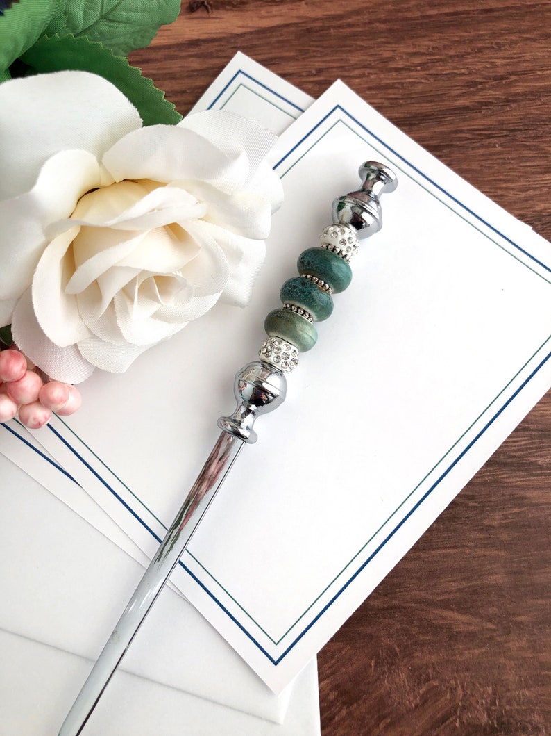 May include: A silver letter opener with a green and white beaded handle. The handle has a silver cap on the top and a silver base at the bottom. The letter opener is lying on top of a stack of white stationery with a blue and green border.