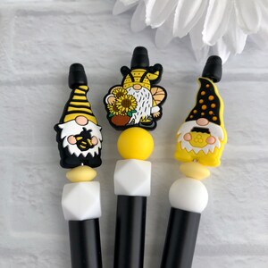 Peut inclure: Trois stylos noirs avec des breloques sur le thème des gnomes jaunes et blancs. Les gnomes portent des chapeaux d'abeilles et tiennent des tournesols.