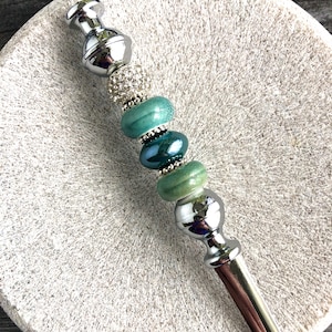 May include: A silver and green beaded hair stick with a decorative top and bottom. The beads are a gradient of green, with a clear crystal bead in the middle.