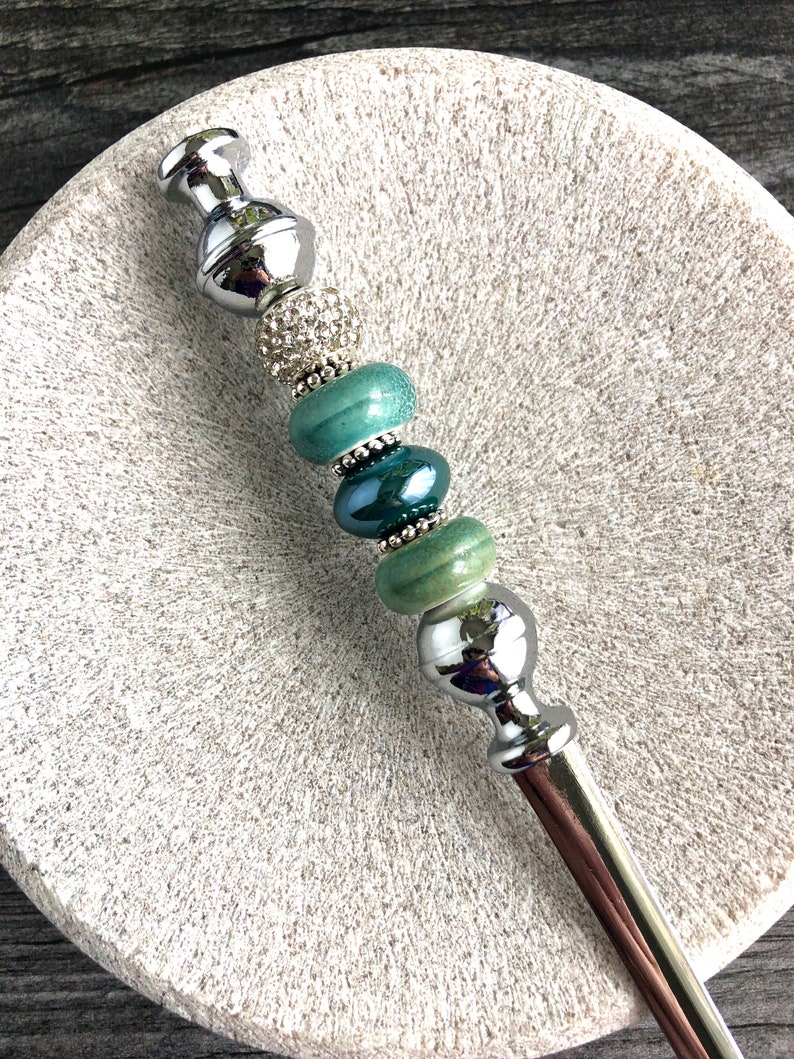 May include: A silver and green beaded hair stick with a decorative top and bottom. The beads are a gradient of green, with a clear crystal bead in the middle.