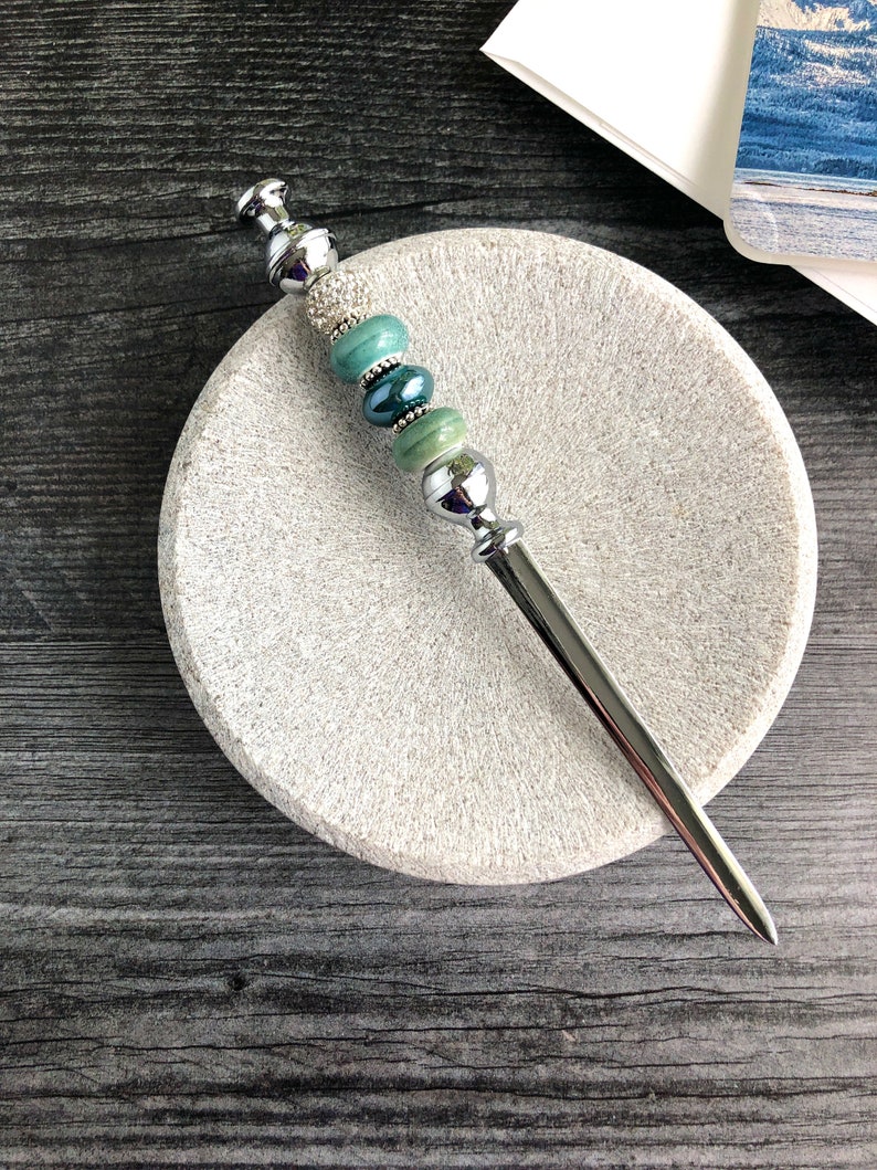May include: A silver letter opener with a decorative handle featuring green and blue beads. The handle is adorned with rhinestones.