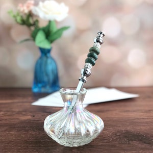 May include: A silver letter opener with green and white beads is placed in a clear glass vase with a textured surface.