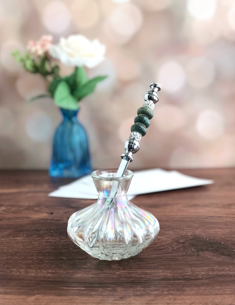May include: A silver letter opener with green and white beads is placed in a clear glass vase with a textured surface.
