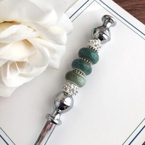 May include: A silver pen with a decorative top featuring a series of green and white beads. The beads are round and have a speckled pattern. The pen is lying on a white surface.