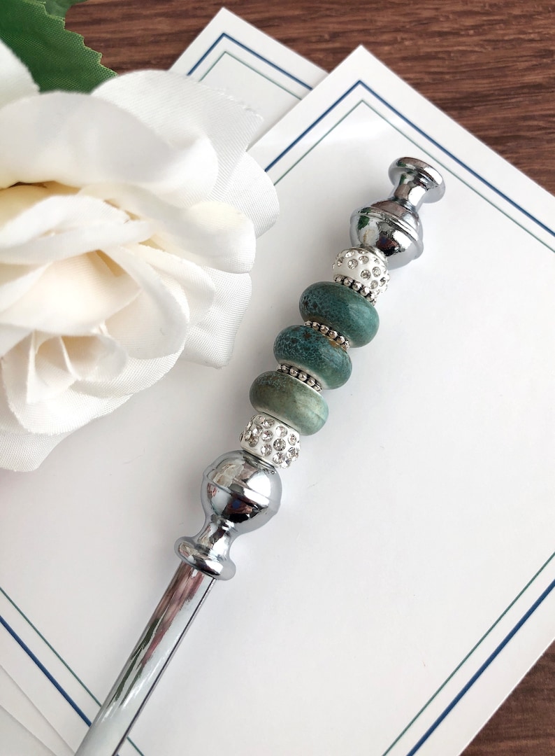 May include: A silver pen with a decorative top featuring a series of green and white beads. The beads are round and have a speckled pattern. The pen is lying on a white surface.
