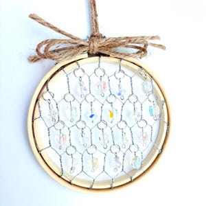 May include: A round, decorative dream catcher with a wooden hoop, wire mesh, and iridescent crystal beads. The dream catcher is suspended by a twine loop and a small bow. The design is simple and elegant.