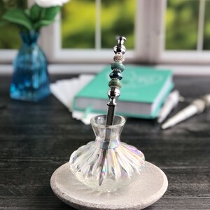 May include: A silver and green beaded pen holder in a clear iridescent glass bottle with a gray stone base.