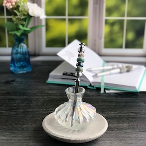 May include: A clear glass pen holder with an iridescent finish, featuring a silver pen with green and blue beads. The pen holder is sitting on a gray stone base.
