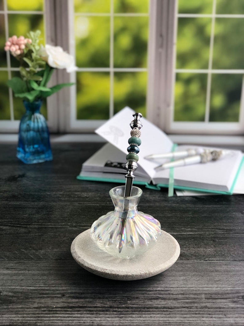 May include: A clear glass pen holder with an iridescent finish, featuring a silver pen with green and blue beads. The pen holder is sitting on a gray stone base.