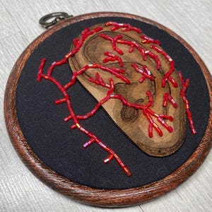 May include: A wooden ear shape is embroidered with red beads on a black fabric background. The embroidery is in the shape of veins or arteries. The fabric is mounted in a dark wood hoop.