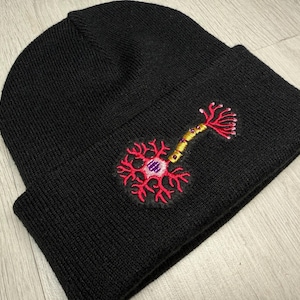Peut inclure: Bonnet en tricot noir avec un bord replié. Le bonnet présente un motif de neurone brodé en rouge, or et rose. Le motif de neurone est sur le devant du bonnet.