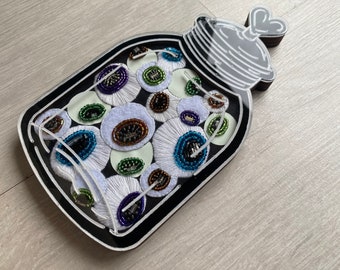 May include: A black wooden jar with a white lid and a heart-shaped handle filled with white, blue, green, and purple embroidered eyeballs.