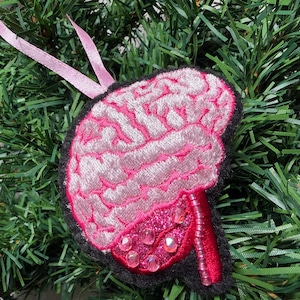 Puede incluir: Adorno en forma de cerebro rosa y blanco con borde negro y cinta rosa para colgar. El cerebro tiene detalles bordados y adornos brillantes. El adorno está sobre un fondo de ramas verdes de árbol de Navidad.