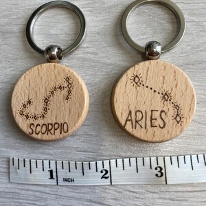 Op de afbeelding: Twee houten sleutelhangers met zilveren sleutelringen. De sleutelhangers zijn rond en hebben de sterrenbeelden Schorpioen en Ram gegraveerd. De sleutelhangers zijn ongeveer 2,5 cm in diameter.