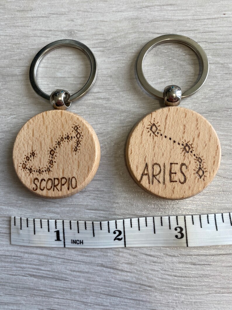 Op de afbeelding: Twee houten sleutelhangers met zilveren sleutelringen. De sleutelhangers zijn rond en hebben de sterrenbeelden Schorpioen en Ram gegraveerd. De sleutelhangers zijn ongeveer 2,5 cm in diameter.