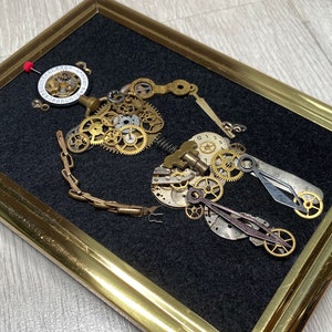 May include: A framed artwork featuring a steampunk-style design made from various clock parts and gears. The artwork is set against a black background and framed in gold.