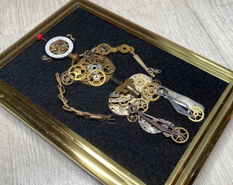 May include: A framed artwork featuring a steampunk-style design made from various clock parts and gears. The artwork is set against a black background and framed in gold.