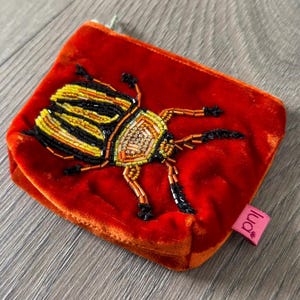 May include: A small, rectangular, red velvet pouch with a beaded insect design. The insect features yellow, black, and gold beads. The pouch has a zipper closure and a small pink tag with the word "lud" on it.