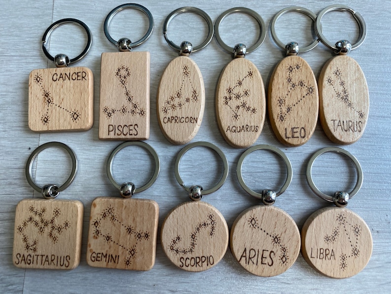 Op de afbeelding: Twaalf houten sleutelhangers met zilveren sleutelringen, elk met een ander sterrenbeeld en de naam ervan. De sterrenbeelden zijn gegraveerd in bruin op het lichtbruine hout. De sterrenbeelden zijn: Kreeft, Vissen, Steenbok, Waterman, Leeuw, Stier, Boogschutter, Tweelingen, Schorpioen, Ram, Weegschaal.