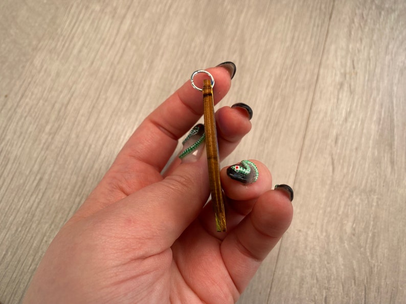 May include: A small, rectangular wooden pendant with a silver-colored ring at the top. The pendant is held in a hand with black nail polish. The wood has a natural, warm brown color.