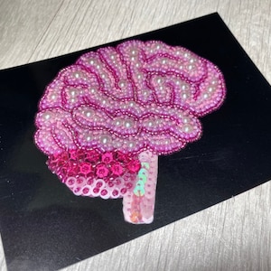 May include: A pink and white beaded brain with a pink and green sequined stem. The brain is on a black background.