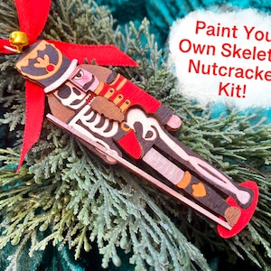 May include: A wooden skeleton nutcracker ornament with a red ribbon and gold bell. The ornament has a skeleton design with red, brown, and white accents. Text on the image reads "Paint Your Own Skeleton Nutcracker Kit!"