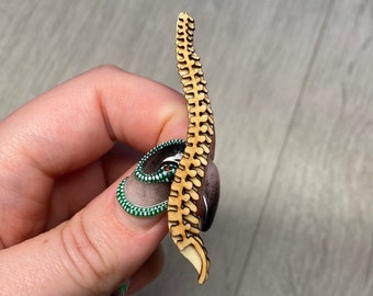 May include: A wooden model of a human spine, with a light brown colour and a detailed design. The spine is held in a hand, with a green and white nail polish on the finger.