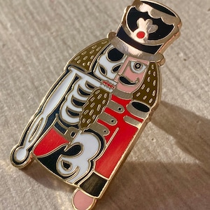 May include: A gold-toned enamel pin featuring a skeleton dressed as a nutcracker. The skeleton is wearing a red and black uniform with a gold trim and a black hat with a red pom-pom. The pin has a detailed design with a white skeleton and a black and gold background.