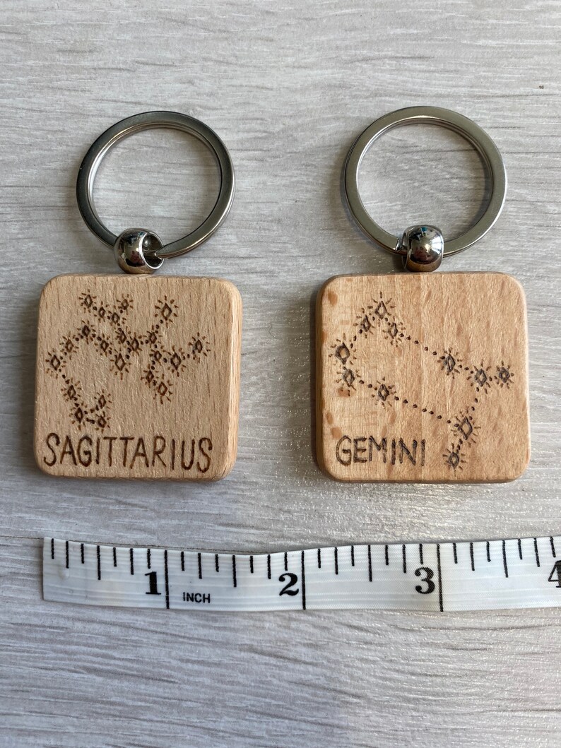 Op de afbeelding: Twee houten sleutelhangers met de sterrenbeelden Boogschutter en Tweelingen, met een liniaal als referentie voor de grootte. De sleutelhangers zijn ongeveer 1 inch vierkant.