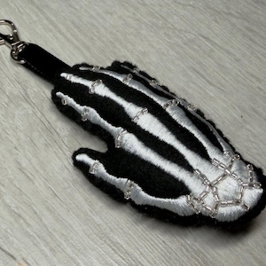 Puede incluir: Llavero de mano de esqueleto en blanco y negro con detalles plateados. El llavero de felpa presenta un diseño de hueso bordado detallado y un cierre plateado para una fácil fijación. Un accesorio único.