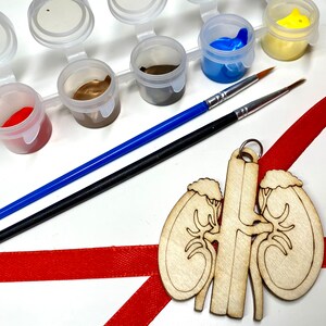 May include: A paint-your-own kit with small paint pots in various colors, two paint brushes, and a wooden kidney-shaped ornament with a red ribbon. The paint colors include red, brown, blue, and yellow.