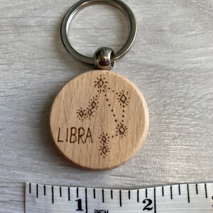 Op de afbeelding: Een houten sleutelhanger met een zilveren sleutelring. De sleutelhanger is rond en heeft het woord "LIBRA" en een sterrenbeeldontwerp gegraveerd. De sleutelhanger is ongeveer 2,5 cm in diameter.