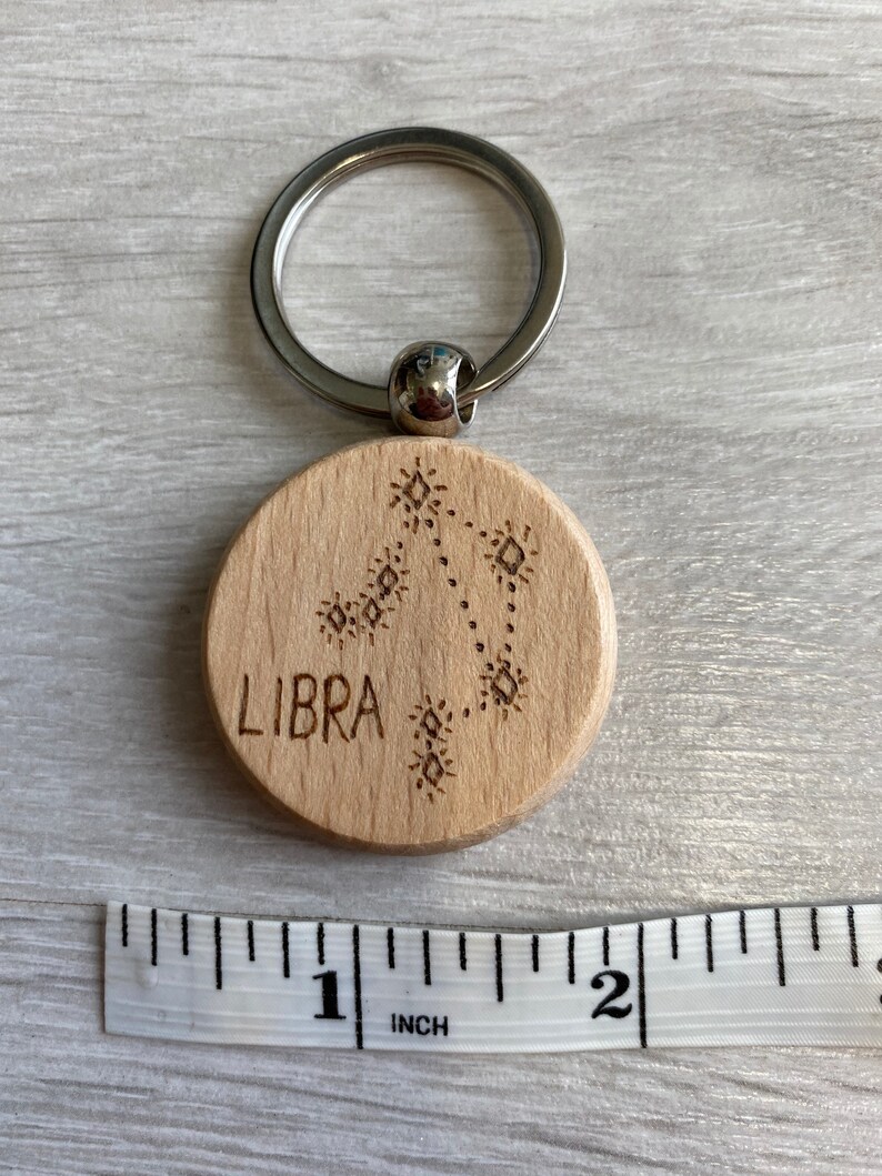 Op de afbeelding: Een houten sleutelhanger met een zilveren sleutelring. De sleutelhanger is rond en heeft het woord "LIBRA" en een sterrenbeeldontwerp gegraveerd. De sleutelhanger is ongeveer 2,5 cm in diameter.