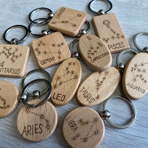 Op de afbeelding: Twaalf houten sleutelhangers met gegraveerde sterrenbeelden en sterrenbeelden. De sleutelhangers zijn in verschillende vormen, waaronder cirkels, ovalen en vierkanten. De tekens omvatten Ram, Stier, Tweelingen, Kreeft, Leeuw, Maagd, Weegschaal, Schorpioen, Boogschutter, Steenbok, Waterman en Vissen.
