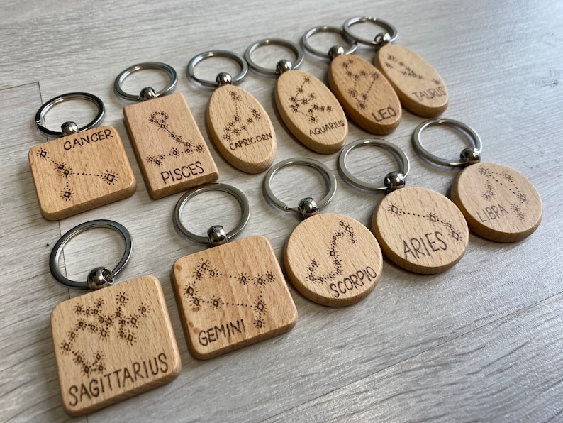 Op de afbeelding: Twaalf houten sleutelhangers met gegraveerde sterrenbeelden. Elke sleutelhanger heeft een ander sterrenbeeld, waaronder Kreeft, Vissen, Steenbok, Waterman, Leeuw, Stier, Weegschaal, Ram, Schorpioen, Tweelingen en Boogschutter.