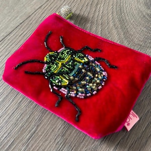 May include: A vibrant fuchsia velvet pouch featuring a beaded beetle design in shades of green, blue, and gold. The beetle is intricately detailed with tiny beads, and the pouch has a small pink tag. The zipper pull is silver.