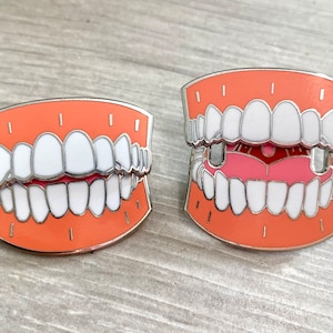Puede incluir: Dos pines de esmalte con forma de dentadura. Los pines son de color naranja y tienen dientes blancos. Un pin tiene una lengua rosa y el otro no.