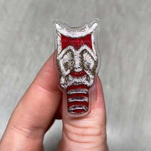 Può includere: Una toppa ricamata rossa e bianca con un design stilizzato di un viso. La toppa è racchiusa in plastica trasparente.