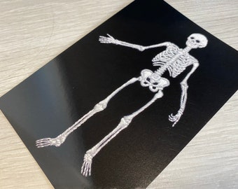 May include: A white skeleton embroidered on a black background. The skeleton is standing with its arms outstretched and its legs spread apart.