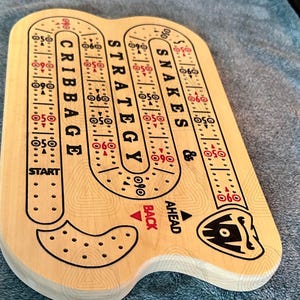 Könnte beinhalten: Ein Holzspielbrett mit einem Schlangen-und-Leitern-Design. Das Brett hat einen gewundenen Pfad mit Zahlen und Pfeilen. Das Brett ist aus hellem Holz und hat ein schwarz-weißes Design. Das Brett ist mit "Snakes & Strategy" und "Cribbage" beschriftet.