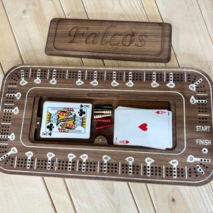 May include: A wooden cribbage board with a lid engraved with the name "Falcos". The board has a recessed area holding playing cards and pegs. The board is brown with white markings and is designed for the game of cribbage.