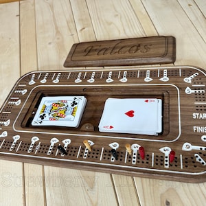 May include: A wooden cribbage board with a lid engraved with "Falcos". The board has a playing surface with holes for pegs, a central card storage area, and includes playing cards and pegs. The board is brown with white accents.