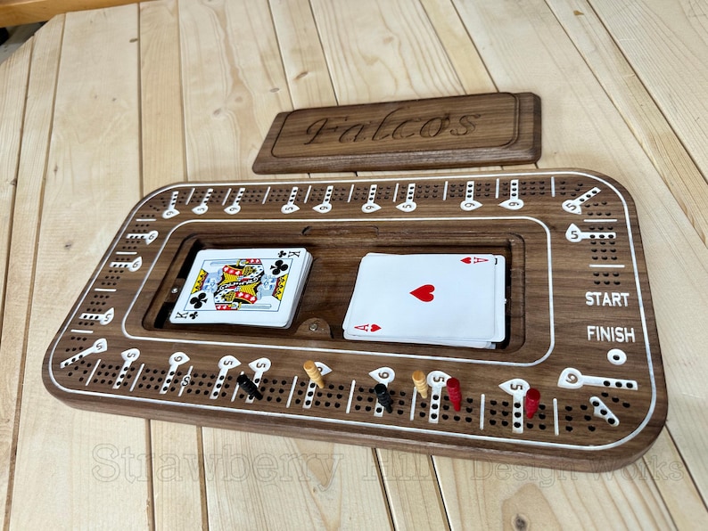 May include: A wooden cribbage board with a lid engraved with "Falcos". The board has a playing surface with holes for pegs, a central card storage area, and includes playing cards and pegs. The board is brown with white accents.