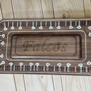 May include: A wooden cribbage board with a rectangular design. The board is engraved with the name "Falcos" and features white markings for scoring. The board has a start and finish area.