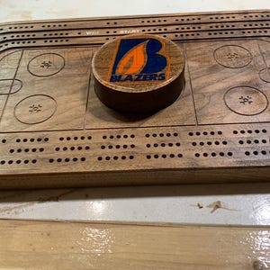 Op de afbeelding: Een houten cribbage-bord met een aangepaste puck met een blauw en oranje logo met de tekst "BLAZERS".