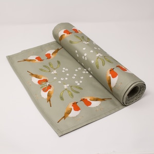 May include: A green table runner with a repeating pattern of red robins, white berries, and green leaves. The runner is rolled up and the pattern is visible on both the rolled and unrolled portions.