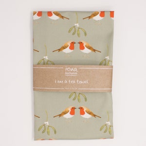 May include: A green tea towel with a repeating pattern of red robins and mistletoe. The towel has a brown label that reads "iona buchanan i am a tea towel made in the uk."