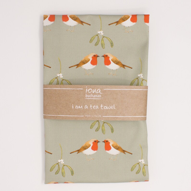 May include: A green tea towel with a repeating pattern of red robins and mistletoe. The towel has a brown label that reads "iona buchanan i am a tea towel made in the uk."