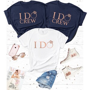 Peut inclure: Trois t-shirts, deux bleus marine et un blanc, avec le texte "I DO CREW" et "I DO" avec un motif d'anneau en or rose. L'image comprend également un short en jean, des lunettes de soleil, un téléphone, un magazine et des baskets blanches.